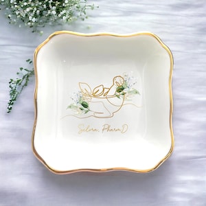 Peut inclure: Un vide-poche carré en céramique blanche avec un bord doré et un motif floral. Le plat a un bord ondulé et présente un dessin au trait doré d'une personne et de fleurs, avec le texte "Selora, Pharm.D" en lettres dorées.
