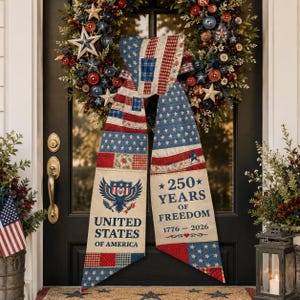 May include: A patriotic door decoration featuring a wreath and a large bow. The bow is made of fabric with a patchwork design of red, white, and blue stars and stripes. The text on the bow reads "United States of America" and "250 Years of Freedom."
