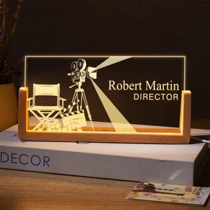 May include: A wooden desk sign with the name "Robert Martin" and the title "DIRECTOR" etched into a clear acrylic panel. The panel features a film camera, director's chair, and clapperboard design. The sign is illuminated.