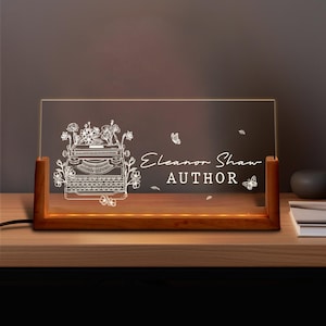 Peut inclure: Lampe de bureau rectangulaire avec une base en bois. Le panneau en acrylique transparent est gravé d'une machine à écrire vintage, de fleurs et du texte "Eleanor Shaw AUTHOR". La base est de couleur marron chaud.