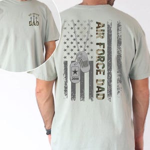 May include: A light green t-shirt with the text "Air Force Dad" and a camouflage American flag design on the back. The front of the shirt has the text "Air Force Dad" with three airplanes above the text.