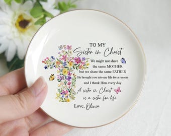 May include: A white ceramic trinket dish with a gold rim. The dish features a floral cross design and the text "TO MY Sister in Christ." Butterflies and additional text are also present. A thoughtful gift.