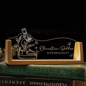 May include: A desk nameplate with a microscope design, illuminated by a warm glow. The clear acrylic plate sits in a wooden base. The name "Christine Robles" and the title "Microbiologist" are displayed in a stylish font.