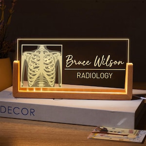 May include: A wooden-based LED light with a rectangular acrylic panel. The panel features a glowing illustration of a human skeleton and the name "Bruce Wilson" with "RADIOLOGY" below. The light is on a book.