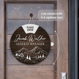 May include: A round, wooden sign with a dark brown finish, featuring the name "Jacob Walker" and the title "General Manager" in white script. The sign includes a mountain and tree design and the text "PLEASE ENTER". It hangs by a rope.