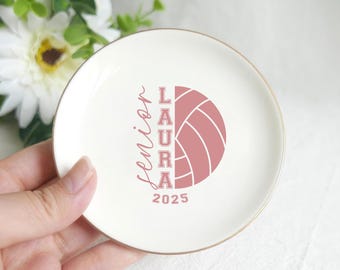 May include: A white ceramic trinket dish with a gold rim. The dish features a pink volleyball graphic and the text "Senior LAURA 2025". The dish is round and appears to be held by a hand.