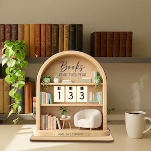 May include: Wooden bookshelf-shaped book counter with the text "Books READ THIS YEAR" and the number "133". The counter has a miniature chair, table, and plants. A mug of coffee sits to the right. The base reads "AMELIA'S LIBRARY".
