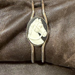 May include: A silver cuff bracelet featuring an oval-shaped cameo centerpiece. The cameo displays a white horse head in profile against a black background, framed by a thin, textured silver border. The bracelet has a split, open-ended band design.