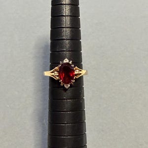 May include: A gold ring with a large oval, deep red gemstone as the centerpiece. The ring has a delicate design with small accent stones on either side of the main stone. The ring is displayed on a black ring sizer.