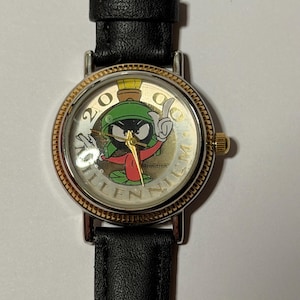 May include: A wristwatch with a black leather band and a gold-toned bezel. The watch face features Marvin the Martian from Looney Tunes, with the text "2000 Millennium" and the Armitron brand name.