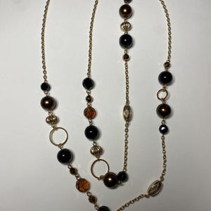 Könnte beinhalten: Eine mehrsträngige Halskette mit einer goldfarbenen Kette und verschiedenen Perlen. Die Perlen sind schwarze, braune und bernsteinfarbene Kugeln, mit dekorativen goldfarbenen Elementen und runden Gliedern. Die Halskette ist lang.