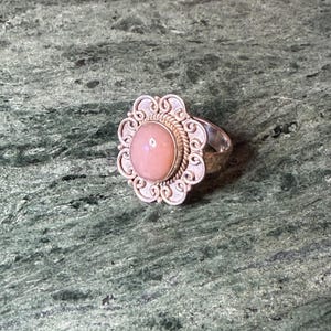 May include: A silver ring with a large, oval, light pink gemstone at the center. The gemstone is surrounded by a decorative, openwork silver setting with a floral design. The ring is set against a green marble surface.