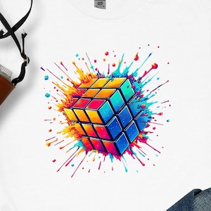May include: White t-shirt featuring a colorful graphic of a Rubik's Cube with a splash effect. The cube has blue, orange, yellow, and red squares. The design is vibrant and eye-catching, perfect for puzzle enthusiasts.