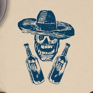 May include: Cream-colored t-shirt featuring a blue skull wearing a sombrero, flanked by two bottles. The design has a woodcut style. The bottles have a circular emblem. The shirt is a casual wear item.