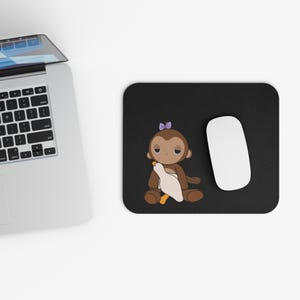 Puede incluir: Alfombrilla de ratón rectangular negra con un mono de dibujos animados con un lazo morado, sosteniendo un ganso blanco con un pico amarillo. Un ratón de ordenador blanco está a la derecha del mono. Un portátil está en la esquina superior izquierda.