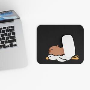 Op de afbeelding: Zwart rechthoekig muismat met een cartoon illustratie van een bruine capibara die op een witte gans rijdt. Een witte computermuis bevindt zich aan de rechterkant van de muismat.