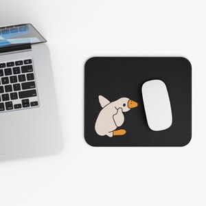 Könnte beinhalten: Ein schwarzes Mauspad mit einem weißen Cartoon-Gänse-Design. Die Gans schaut nach rechts und hat ihren Flügel erhoben. Eine weiße Computermaus befindet sich auf dem Mauspad.