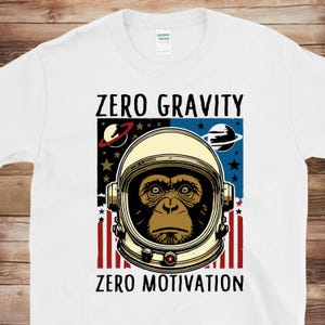 May include: White t-shirt featuring a graphic of a monkey wearing an astronaut helmet. The design includes the text "ZERO GRAVITY" above the helmet and "ZERO MOTIVATION" below. The background has a space theme with planets and stars.