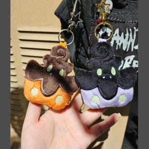 May include: Two plush keychains in the shape of a cupcake or ghost. One is brown and orange, the other is black and purple. Both have green embroidered details and gold key rings.