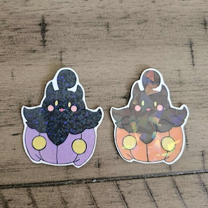 May include: Two stickers featuring a cartoon character with a pumpkin-shaped body and bat-like wings. One has a purple body and black glittery wings, the other has an orange body and brown wings. Both have yellow circles and pink cheeks.