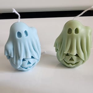 May include: Two ghost-shaped candles. One is light blue, the other is green. Each candle has a carved pumpkin face at the bottom and a draped sheet-like form for the ghost's body. White wicks are visible.