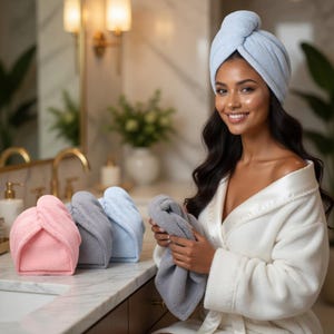 Pode incluir: Uma mulher usando um turbante de toalha azul claro, segurando uma toalha cinza. Três turbantes de toalha adicionais em rosa, cinza e azul claro estão em uma bancada de mármore. A mulher está vestindo um roupão branco.
