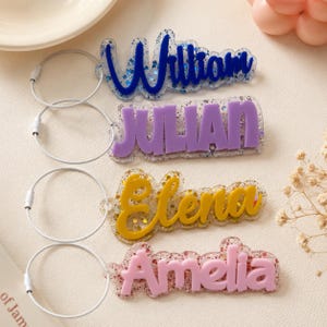 Può includere: Quattro portachiavi personalizzati con nomi. I nomi sono William in blu, Julian in viola, Elena in giallo e Amelia in rosa. Ogni nome è attaccato a un anello di metallo color argento.