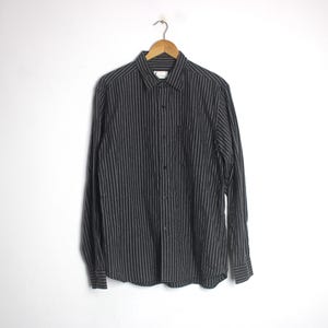 May include: A long-sleeve button-down shirt in a dark charcoal gray fabric featuring thin, vertical white pinstripes. The shirt includes a standard collar, a front button placket, and buttoned cuffs, displayed hanging on a wooden hanger.