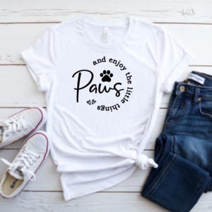 May include: White t-shirt with the text "Paws and enjoy the little things" in a circular design with a paw print. The shirt is paired with blue jeans and white sneakers with red accents, all on a light wood surface.