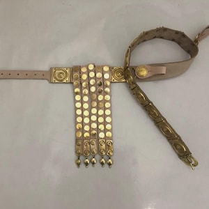 May include: A beige leather wrist cuff with gold-colored metal accents. The cuff features a series of gold discs and decorative squares. A matching beige leather strap and a gold-colored metal arm band are also included.