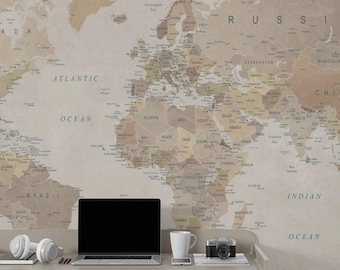 May include: A workspace featuring a world map wallpaper in shades of beige and brown. A laptop with a blank screen, headphones, a coffee mug, a smartphone, and a camera are on a desk. A chair is in the foreground.