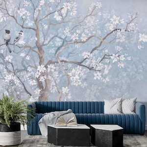 May include: A blue velvet sofa set against a wall mural of a tree with white blossoms and two birds. The room features a potted plant, two black and white coffee tables, and decorative cushions.