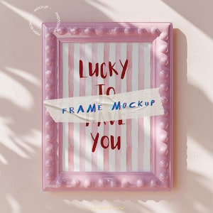 May include: A pink rectangular frame with a beaded edge. Inside the frame is a print with red and white vertical stripes and the words "LUCKY TO HAVE YOU" in red. A piece of tape with the words "FRAME MOCKUP" is across the print.