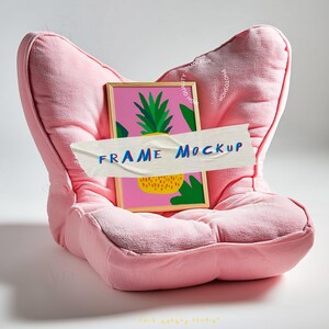 May include: A pink, plush armchair with a framed print featuring a pineapple design. The print is covered by a piece of tape with the words "FRAME MOCKUP" in blue. The frame is made of wood.
