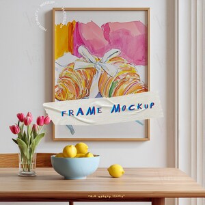 May include: A framed watercolour print of two croissants tied with a white ribbon. The artwork has a light brown frame and is taped with a banner that reads "FRAME MOCKUP". A vase of pink tulips, a blue bowl of lemons, and a single lemon are on a wooden table.