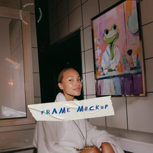 May include: A framed artwork featuring a frog in a white robe, with makeup products in front of a mirror. The artwork is in a gold frame. The words "FRAME MOCKUP" are on a piece of tape in the foreground.