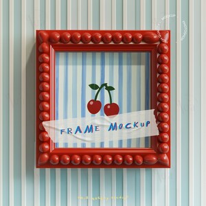 Op de afbeelding: Een rode vierkante lijst met een kralenrand, met een kersenillustratie op een gestreepte achtergrond. De woorden "FRAME MOCKUP" zijn zichtbaar op een stuk tape. De lijst is gemonteerd op een blauw-wit gestreepte muur.