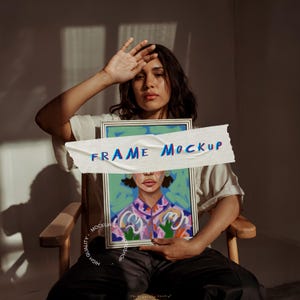 Puede incluir: Una persona sostiene un cuadro enmarcado de una mujer con ropa colorida sobre un fondo azul. El marco está parcialmente cubierto por un trozo de papel blanco con el texto "FRAME MOCKUP" en letras azules y rojas.