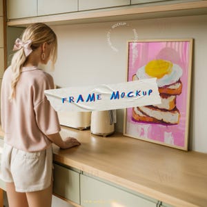 Puede incluir: Una impresión artística enmarcada con una pintura colorida de un huevo frito sobre tostadas. La obra de arte tiene un fondo rosa y se muestra en un marco de madera clara. Las palabras "FRAME MOCKUP" están en una cinta adhesiva frente al arte.
