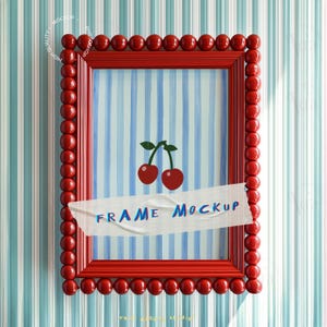 Puede incluir: Un marco de fotos rojo con un borde de cuentas y una abertura rectangular. El marco contiene una pintura de cerezas y rayas azules y blancas. El texto "FRAME MOCKUP" está en un trozo de cinta adhesiva.