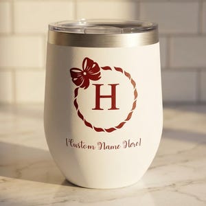 Pode incluir: Um copo de vinho branco sem haste com borda prateada e tampa transparente. O copo apresenta um laço vermelho e um desenho de coroa em volta da letra "H". Abaixo da letra está o texto "Custom Name Here!"