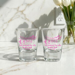 May include: Two clear glass shot glasses featuring pink printed text that reads a bombshell bride has entered the villa, Sarah's Bach, and Cancun 2026. The design includes a string of icons depicting a high heel, ring, bikini, and star.