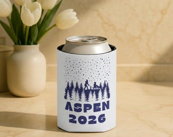 May include: White can cooler with a blue graphic of a forest, people, and falling snow. The text "ASPEN 2026" is printed in blue. The cooler is holding a silver can.