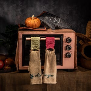 Puede incluir: Un horno tostador rosa con una peque&ntilde;a calabaza naranja encima, junto a un guante de horno negro con texto blanco. Dos pa&ntilde;os de cocina beige cuelgan del horno, cada uno con una parte superior de ganchillo y el texto "olive you".