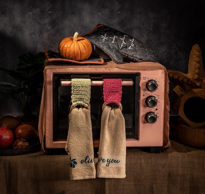 Puede incluir: Un horno tostador rosa con una peque&ntilde;a calabaza naranja encima, junto a un guante de horno negro con texto blanco. Dos pa&ntilde;os de cocina beige cuelgan del horno, cada uno con una parte superior de ganchillo y el texto "olive you".
