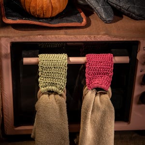 Puede incluir: Una naturaleza muerta con una peque&ntilde;a calabaza naranja, un guante de horno negro con texto blanco y dos pa&ntilde;os de cocina colgantes. Las toallas son beige con soportes de ganchillo verdes y rojos. El texto "olive you" es visible.