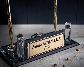 Puede incluir: Un juego de escritorio de cristal negro con dos bolígrafos dorados, dos pilares de cristal transparente y una placa central de metal dorado con el texto Name SURNAME Title sobre una base rectangular de cristal negro reflectante.