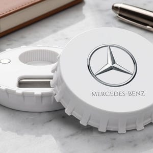 Puede incluir: Un abridor de botellas circular blanco con un borde dentado que recuerda a una chapa. La superficie superior presenta el logotipo plateado de Mercedes-Benz y el texto MERCEDES-BENZ. El dispositivo tiene una abertura central y mecanismo.