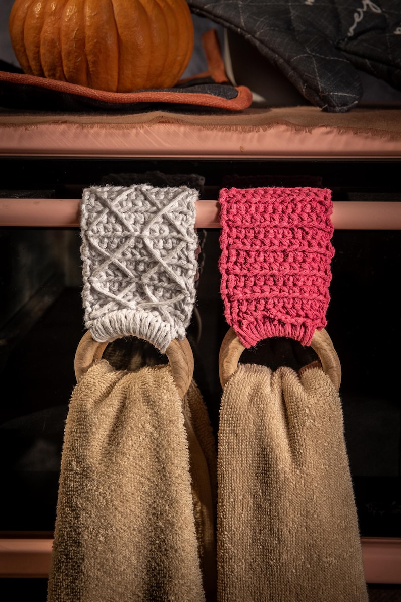 Puede incluir: Dos soportes para pa&ntilde;os de cocina con aros de madera y toallas beige. Un soporte es gris con un patr&oacute;n de diamantes, el otro es rosa brillante. Una calabaza y manoplas para horno est&aacute;n en el fondo.