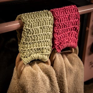 Puede incluir: Dos pa&ntilde;os de cocina colgados de una barra. Las toallas son beige y tienen aros de madera. Los soportes est&aacute;n tejidos a ganchillo, uno en color verde claro y otro en color rosa brillante.